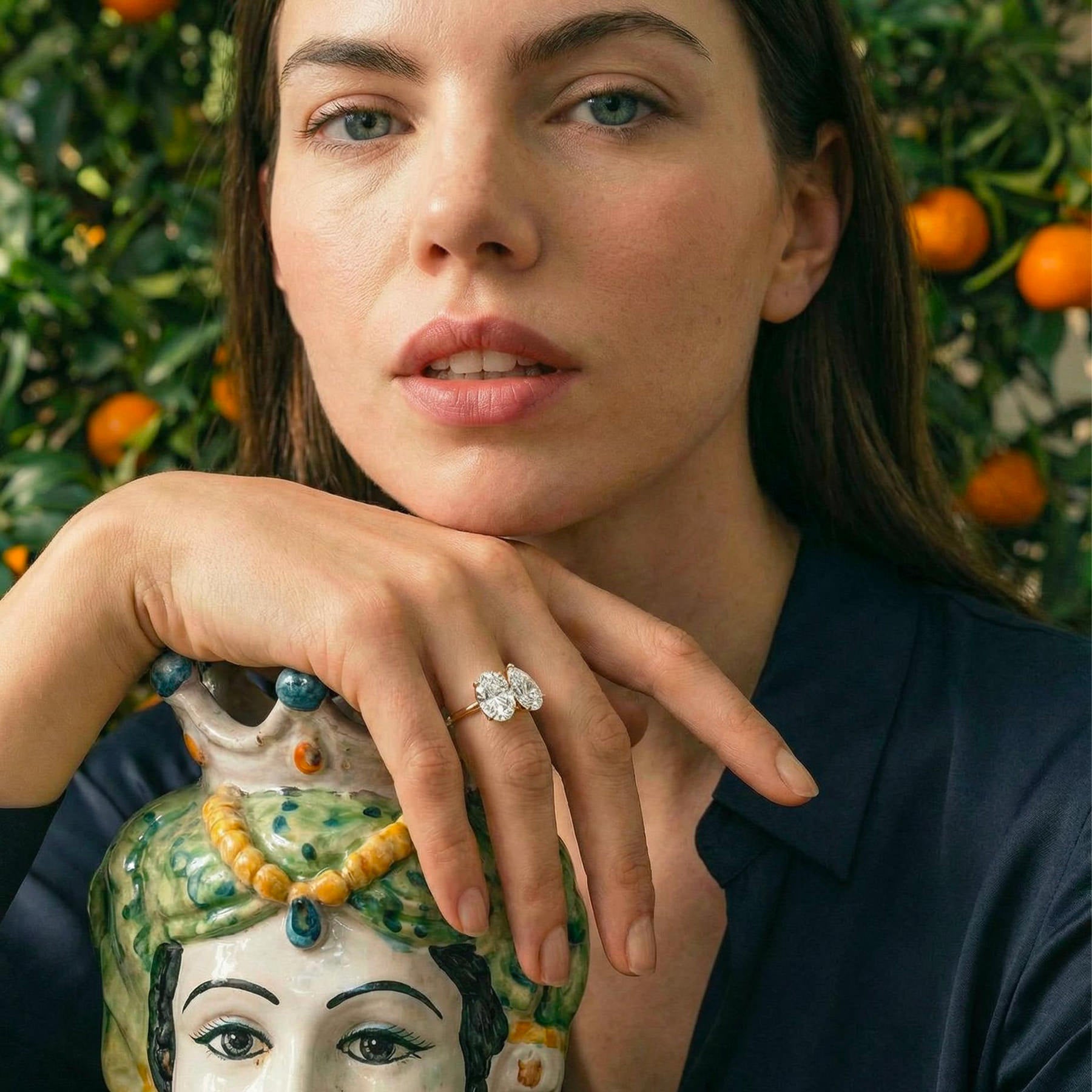 Bague de fiançailles Toi et Moi Maison Mandarine en or 18 carats avec diamants taille ovale et poire portée par une femme devant un mandarinier avec la main posée sur un vase sicilien testa di moro